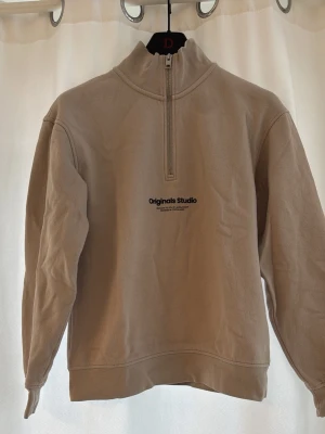 Beige halfzip från Jack & Jones - Säljer en beige sweatshirt med halv dragkedja från Jack & Jones. Tröjan har hög krage, tryckt logga på bröstet och är tillverkad i mjukt bomullsmaterial. Perfekt för en avslappnad och stilren look.