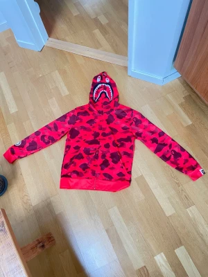 Röd BAPE Shark Hoodie med camomönster - Säljer en röd BAPE Shark Hoodie med ikoniskt camomönster och hajmotiv på huvan. Dragkedja framtill och detaljerad logga på ärmen. Perfekt för dig som vill sticka ut med streetwear från A Bathing Ape. Materialet är mjukt och bekvämt, troligen bomullsblandning.