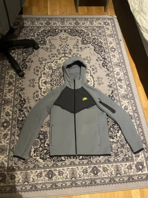 Grå Nike Tech Fleece hoodie - Snygg grå Nike Tech Fleece hoodie med svart panel över bröstet och gul Nike-logga. Dragkedja framtill, fickor på sidorna och en extra ficka på ärmen. Tillverkad i mjukt och stretchigt material, perfekt för en sportig och avslappnad stil.