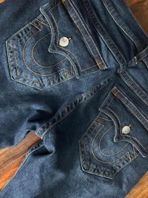 Mörkblå True Religion jeans - Säljer ett par mörkblå jeans från True Religion med ikoniska kontrastsömmar och stora fickor bak med dekorativa knappar. Klassisk rak passform och tillverkade i kraftigt denimtyg. Perfekta för dig som gillar streetwear och vill sticka ut med detaljer.