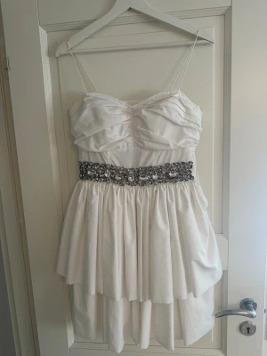 Vit kort klänning med paljettbälte - Supersöt vit kort klänning med tunna axelband och volangdetaljer. Midjan pryds av ett brett bälte med glittrande paljetter och pärlor som ger extra wow-känsla. Kjolen är dubbellager och ger en fluffig look. Ser som ny ut säljes pågrund av ingen användning. 
