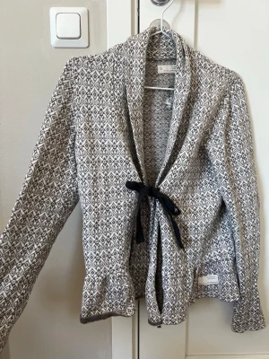 Odd Molly mönstrad kofta med knytband - Supersnygg kofta från Odd Molly med gråvitt grafiskt mönster och svart knytband framtill. Koftan har långa ärmar, volangdetaljer vid ärmslut och nederkant. Står ingen storlek på den men skulle säga att den passar S/M💕