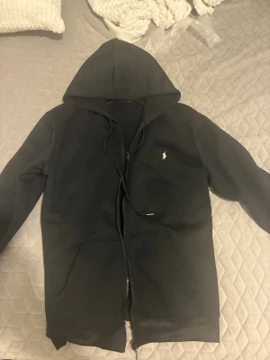 Svart hoodie från Polo Ralph Lauren - Klassisk svart hoodie från Polo Ralph Lauren med dragkedja framtill och vit broderad logga på bröstet. Hoodien har huva med dragsko och två fickor framtill. Tillverkad i mjukt bomullsmaterial som känns skönt mot huden.