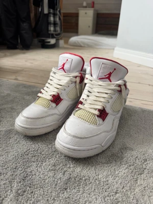 Air Jordan 4 Retro Vit/Röd - Säljer ett par Air Jordan 4 Retro sneakers i vitt läder med röda detaljer och meshpaneler. Skorna har den klassiska Jumpman-loggan på plösen och vita skosnören. Snygg siluett med högt skaft och ikoniska plastdetaljer vid snörningen.