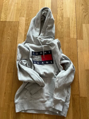 Grå hoodie från Tommy Jeans - Snygg grå hoodie från Tommy Jeans med stor logga i blått, vitt och rött på bröstet. Klassisk känguruficka framtill och justerbar huva med dragsko. Tillverkad i mjukt sweatshirtmaterial som är perfekt för chill dagar.