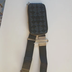 Svart Givenchy handväska med mönster - Säljer en svart handväska från Givenchy med präglat geometriskt mönster och silverfärgad dragkedja. Väskan har en bred axelrem med metallspänne och Givenchy-logga på remmen. Perfekt för dig som gillar stilrena accessoarer med edge. Nypris 12000