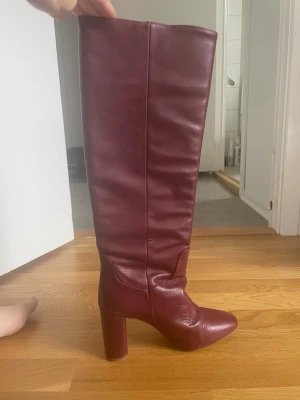Vinröda knähöga boots med klack - Säljer ett par vinröda knähöga boots i slätt skinnimitation med rundad tå och chunky blockklack. Stövlarna har en stilren design utan synliga detaljer och ger en cool retrovibe. Perfekta för att lyfta vilken outfit som helst.