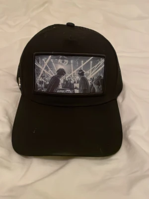 Svart truckerkeps med DJ-motiv - Säljer en svart Maggior justerbart spänne. Framtill finns ett stort tryck med DJ-motiv och klubbkänsla i svartvitt. Perfekt för dig som gillar musik och streetwear. Kepsen har böjd skärm och klassisk passform.