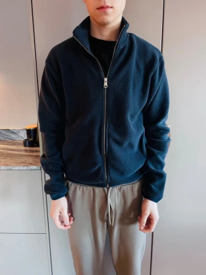 Marinblå fleecejacka J.Lindeberg - Mysig marinblå fleecejacka från J.Lindeberg med dragkedja framtill och två fickor. Hög krage och enkel, stilren design som passar perfekt till lager-på-lager. Tillverkad i mjukt fleecematerial för extra komfort.