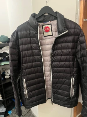 Svart dunjacka från Colmar - Snygg svart dunjacka från Colmar med quiltad design och silvriga dragkedjor. Jackan har ståkrage, två fickor med dragkedja och lätt vadderad insida i ljusgrått. Perfekt för kalla dagar när du vill hålla stilen.