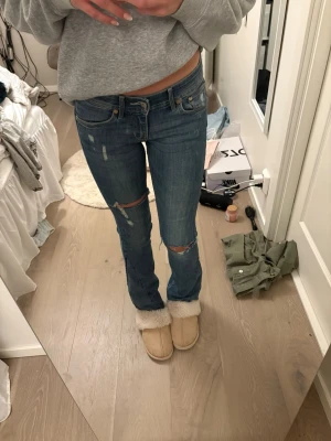 Blå slitna bootcut jeans - Säljer ett par blå bootcut jeans med slitningar och hål på knäna. Jeansen har låg midja och en snygg tvättad look. Perfekta för en avslappnad och trendig stil.