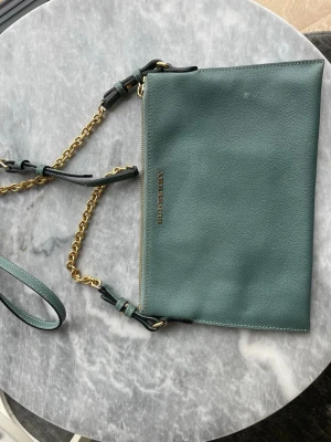 Grön axelväska från Burberry - Snygg grön axelväska från Burberry i läder med guldfärgad kedja och justerbar axelrem. Väskan har en stilren, rektangulär form och diskret Burberry-logga i guld på framsidan. Perfekt för dig som gillar klassiska designer och lyxiga detaljer. En fläck på framsidan som jag inte får bort. Rutigt Burberry mönster på insidan med 4 st fack för kort. Fin men använd. Mått: 25,5 x 17 cm