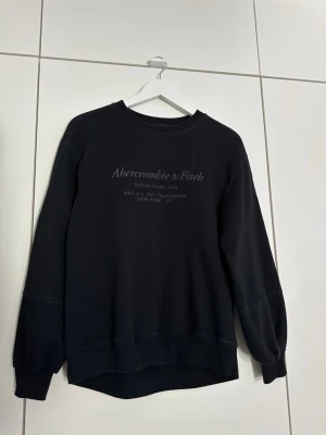 Svart sweatshirt från Abercrombie & Fitch - Svart sweatshirt från Abercrombie & Fitch med diskret logga på bröstet. Tröjan har rund hals, långa ärmar och ribbade muddar. Materialet är mjukt och bekvämt, perfekt för en avslappnad look. Passar dig som gillar klassisk och stilren design.