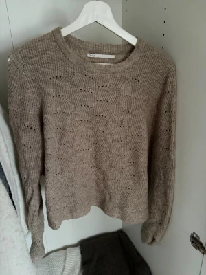 Beige stickad tröja från ONLY - Stickad tröja i beige från ONLY med rund halsringning och ett diskret vågmönster i strukturen. Tröjan har långa ärmar och en avslappnad passform, perfekt för lager på lager under kyliga dagar. Materialet ser ut att vara mjukt och bekvämt.