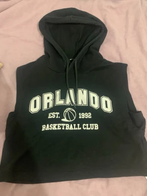 Svart Orlando hoodie utan ärmar - Cool svart hoodie utan ärmar med trycket 'ORLANDO BASKETBALL CLUB EST. 1992' i vitt framtill. Har en stor huva med dragsko och sportig vibe. Perfekt för dig som gillar basketstil och vill ha något avslappnat men ändå trendigt.