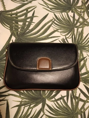 Svart och brun läderhandväska - Snygg svart handväska i äkta slätt läder (ko skinn) med bruna detaljer och kontrastsömmar. Väskan har ett lock med magnetknäppning och en innerficka med dragkedja. Superfin och helt ny. Skriv privat vid frågor☺️