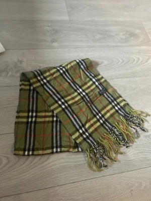 Rutig ullscarf från Burberrys - Säljer en klassisk rutig scarf från Burberrys i 100% lammull. Scarfen har en grön bas med inslag av svart, vitt, beige och röda linjer. Tidlös design med fransar i nederkant och mjuk ullkänsla. Perfekt för dig som vill ha en ikonisk look.
