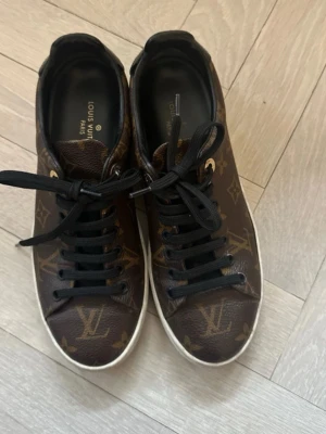 Louis Vuitton monogram sneakers - Snygga sneakers från Louis Vuitton i klassiskt brunt monogrammönster med svarta skosnören och vita sulor. Skorna har guldfärgade detaljer vid snörningen och logotyp på sidan av sulan. Tillverkade i läder med lyxig känsla och ikonisk design.