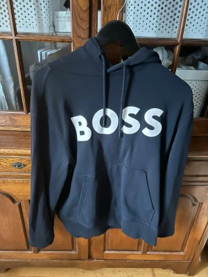 Marinblå hoodie från BOSS med logga - Säljer en marinblå hoodie från BOSS med stor vit logga på bröstet. Hoodien har huva med dragsko och en stor magficka. Perfekt för dig som gillar stilrena och klassiska plagg. Materialet, bomull känns mjukt och bekvämt. 
