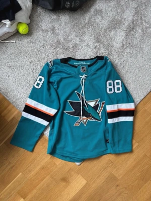 San Jose Sharks NHL matchtröja #88 - Säljer en tealgrön San Jose Sharks hockeytröja med nummer 88 på ärmarna. Tröjan har snörning i halsen, stora loggan på bröstet och detaljer i svart, vitt och orange på ärmarna. Perfekt för dig som älskar hockey och vill representera ditt lag.