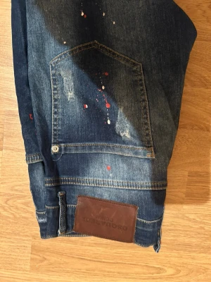 Desigual jeans med färgstänk - Unika blå jeans från Desigual med coola färgstänk i rött och vitt, slitningar och broderade detaljer. Klassisk femficksmodell med läderpatch bak. Perfekt för dig som gillar en kreativ och edgy stil.