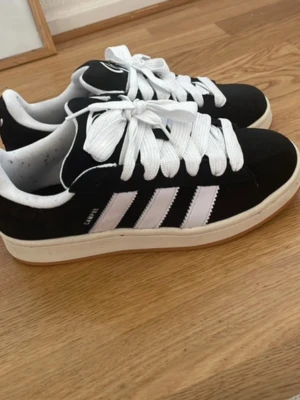 Adidas Campus svartvita sneakers - Säljer ett par klassiska Adidas Campus sneakers i svart mocka med vita detaljer och breda vita skosnören. Skorna har de ikoniska tre ränderna på sidan och en vit sula. Perfekta för en clean och sportig stil.