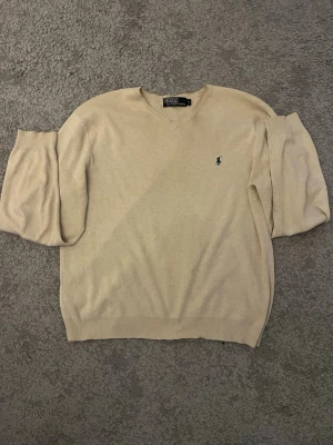 Beige stickad tröja Polo Ralph Lauren - Snygg beige stickad tröja från Polo Ralph Lauren med klassisk v-ringning och den ikoniska gröna loggan broderad på bröstet. Tröjan har ribbade muddar vid ärmslut och nederkant. Perfekt för dig som gillar stilrena och tidlösa plagg. Fläckar förekommer men bör enkelt gå bort! Sen är det en liten söm i v ringen som släppt de bild 2