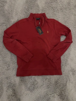 Röd ziptröja från Polo Ralph Lauren - Snygg röd ziptröja från Polo Ralph Lauren med hög krage och dragkedja. Tröjan har det klassiska broderade logot på bröstet och ribbade muddar vid ärmar och nederkant. Perfekt för en avslappnad men stilren look. Tröjan är L i barnstorlek vilket är Xs i herr storlek 