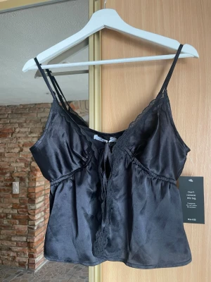 Svart satinlinnetopp med spets detaljer - Snygg svart linnetopp från NA-KD i glansigt satinmaterial med tunna axelband och spetsdetaljer längs ringningen. Toppen har en liten knytning framtill. Aldrig använd 🌸