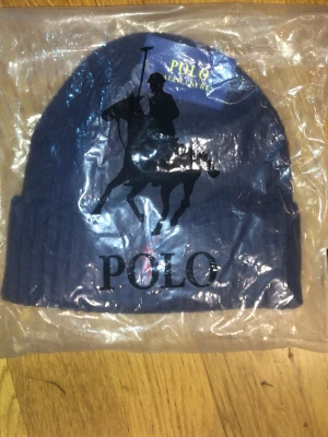 Mörkblå mössa från Polo Ralph Lauren - Snygg mörkblå ribbstickad mössa från Polo Ralph Lauren med klassisk broderad röd logga på uppviket och stor svart polospelare tryckt framtill. Perfekt accessoar för att hålla värmen och addera stilpoäng till din outfit.