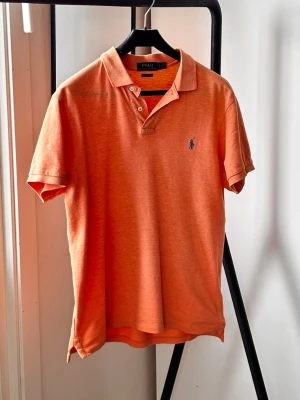 Pikétröja från Polo Ralph Lauren - Säljer en orange pikétröja från Polo Ralph Lauren i slim fit-modell. Tröjan har klassisk krage, korta ärmar och knappar framtill. Tillverkad i mjuk bomull och har den ikoniska broderade loggan på bröstet.