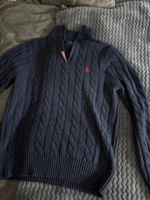 Mörkblå kabelstickad tröja Polo Ralph Lauren - Snygg mörkblå kabelstickad tröja från Polo Ralph Lauren med halv dragkedja och ståkrage. Tröjan har det klassiska röda Polo-logot på bröstet och ribbade muddar vid ärmslut och nederkant. Perfekt för lager på lager och en clean look.