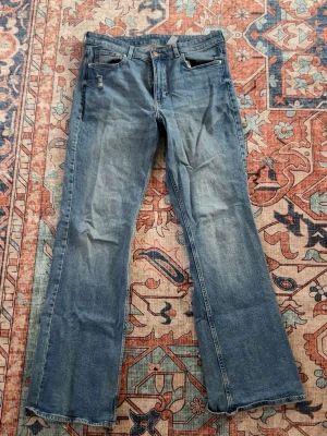 Blå bootcut jeans med rå kant - Säljer ett par blå bootcut jeans med klassisk femficksmodell och rå, fransad kant längst ner på benen. Jeansen har en snygg tvätt med slitningar framtill och är tillverkade i denim. Perfekta för en avslappnad och trendig look. Lite slitna där nere.