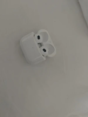 Trådlösa vita hörlurar med laddetui - Säljer ett par trådlösa vita hörlurar med matchande laddningsetui. Hörlurarna har en stilren och kompakt design med rundade kanter och svarta detaljer på insidan. Perfekta för musik, poddar eller samtal på språng. Materialet är blank plast. ( AirPod 3)