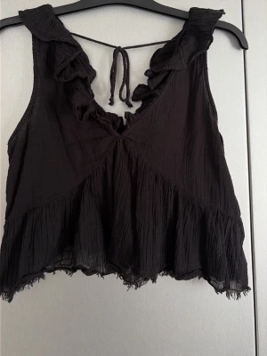 Svart volang topp från Gina Tricot - Snygg svart ärmlös topp från Gina Tricot med volangdetaljer längs urringningen och nertill. Toppen har knyt i nacken och är tillverkad i ett tunt, lätt material som ger en luftig känsla. Perfekt för att lyfta vilken outfit som helst. Storlek m men är liten i storlek! 