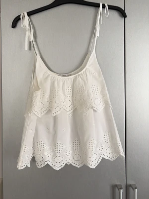 Vit broderad linnetopp från Pigalle - Söt vit linnetopp från Pigalle med broderade spetsdetaljer och volanger. Toppen har tunna axelband med knyt och en luftig, flowy passform. Perfekt för varma dagar och ger en romantisk vibe till din outfit. Endast använd en gång!