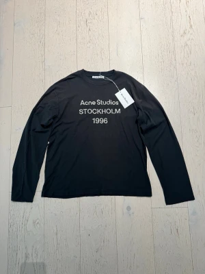 Svart långärmad top Acne Studios - Svart långärmad topp från Acne Studios med vit text 'Acne Studios STOCKHOLM 1996' på bröstet. Klassisk rund halsringning och rak passform. Perfekt för dig som gillar minimalistisk och stilren design.