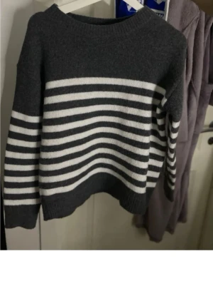 Mörkgrå stickad tröja med ränder - Snygg mörkgrå stickad tröja med vita horisontella ränder över bröstet och ärmarna. Tröjan har rund halsringning och ribbstickade muddar. Perfekt för en avslappnad och trendig look.