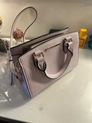 ALDO Ljuslila/rosa handväska med axelrem - Så fin ljuslila/rosa väska från ALDO i strukturerat material med gulddetaljer.  Kan bäras både i handen och som crossbody tack vare det långa justerbara axelbandet. Stängs med dragkedja och har två innerfickor som gör den praktisk trots sin smidiga storlek.  Superfint skick och väldigt sparsamt använd! Perfekt till både vardag, fest eller sommaroutfits. 