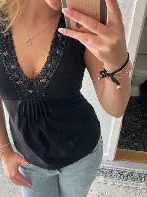 Svart ärmlös topp med spetsdetaljer - Snygg svart ärmlös topp med djup v-ringning och spetskant dekorerad med små blommor. Toppen har en lätt rynkad detalj under bysten och är gjord i ett mjukt material som känns bekvämt mot huden. Perfekt att styla med jeans eller kjol. Okänt märke, köptes av en annan tjej på loppis. Storlek XS/S 