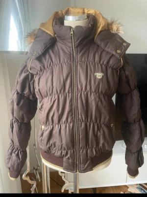 Brun pufferjacka ny  - Säljer en brun pufferjacka från JLO med guldfärgade detaljer och avtagbar huva med fuskpäls. Jackan har dragkedja framtill, två fickor med tryckknappar och ribbade muddar 
