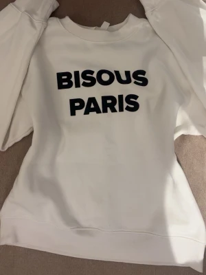 Vit sweatshirt med text Bisous Paris - Säljer en vit sweatshirt med svart text 'BISOUS PARIS' på. Tröjan har rund halsringning och långa ärmar. Materialet är mjukt och bekvämt mot huden.