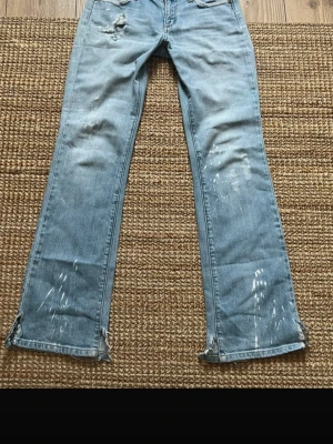 Ljusblå bootcut jeans med slitningar - Säljer ett par ljusblå bootcut jeans med slitningar och detaljerade revor på både fram- och baksida. Jeansen har en cool, avslappnad look. Perfekta för dig som gillar en trendig och lite ruff stil.