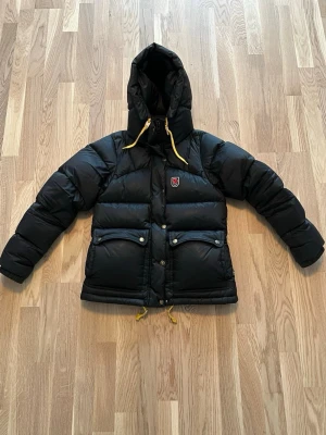 Svart pufferjacka från Fjällräven - Snygg svart pufferjacka från Fjällräven med huva och gula dragskor. Jackan har två stora fickor med lock och tryckknappar framtill samt Fjällräven-logga på bröstet. Perfekt för kalla vinterdagar och riktigt skön att bära. Klassisk design och rejält vadderad.