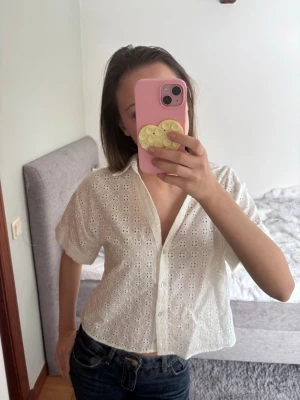 Blus - Söt vit blus med broderade hålmönster och korta ärmar. Blusen har knappar framtill och en avslappnad, boxig passform. Perfekt till jeans eller kjol för en fräsch look. Materialet känns lätt och luftigt, perfekt för varmare dagar.