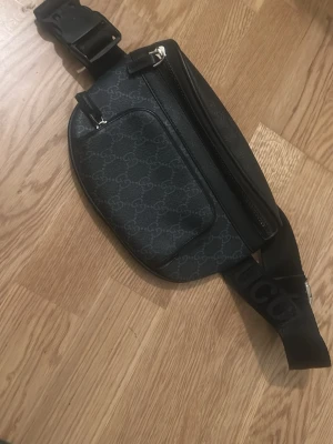 Svart magväska från Gucci - Snygg svart magväska från Gucci med klassiskt GG-mönster i grått. Väskan har två dragkedjefack och justerbart band med Gucci-logga. Tillverkad i slitstarkt material med detaljer i läder och metall. Perfekt för att bära det viktigaste på ett stilrent sätt.