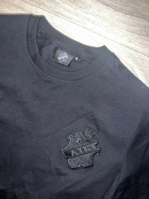 Svart AIK t-shirt  - Svart AIK t-shirt oanvänd med en broderad logotyp på bröstet. T-shirten har en kontrasterande kant i en ljusare färg (guld,beige), vilket ger en stilren detalj. Halsringningen är rund och ribbad.