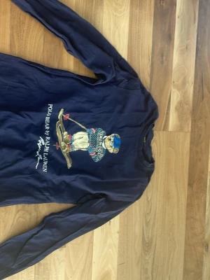 Marinblå långärmad t-shirt Polo Bear - Marinblå långärmad t-shirt från Ralph Lauren med Polo Bear-motiv på bröstet. Trycket visar en björn i stickad tröja och skidutrustning. T-shirten har rund hals och är tillverkad i bomull. Perfekt för dig som gillar klassiska och ikoniska prints.