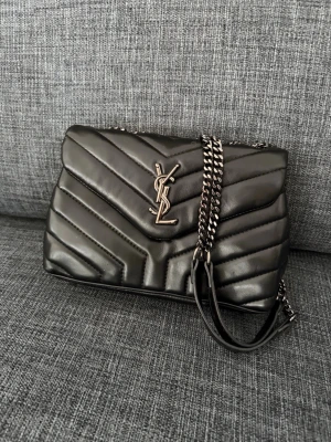 Svart YSL handväska i läder - Säljer en svart quiltad handväska från Saint Laurent med ikoniskt YSL-emblem i silver framtill. Väskan har kedjeremmar i silverfärg och är tillverkad i mjukt läder med elegant mönster. Perfekt accessoar för dig som gillar lyxig och tidlös design. Fick den av gammelmormor när hon gick bort 