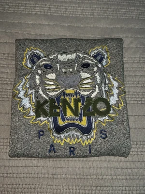 Grå Kenzo sweatshirt med tiger - Säljer en grå sweatshirt från Kenzo med ikonisk broderad tiger i vitt, blått och gult på bröstet samt texten 'KENZO PARIS'. Tröjan har rund halsringning och ribbade muddar. Perfekt för dig som gillar streetwear och vill sticka ut.
